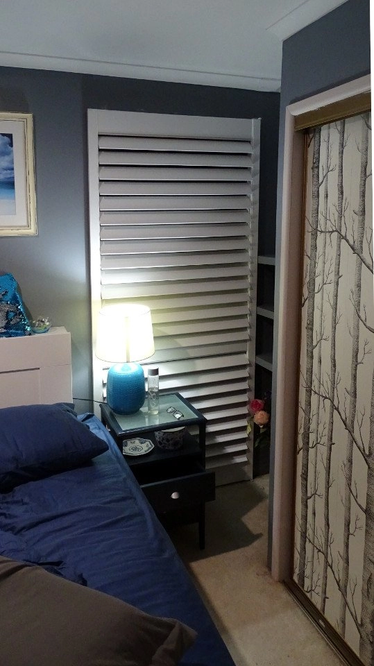 Plantation shutters installed on a window next to a bedside table with a lamp and decorative items.