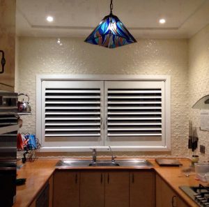 White Plantation Shutters in Kitchen Window 1 White plantation shutters installed in a kitchen window above a sink with a colorful pendant light.